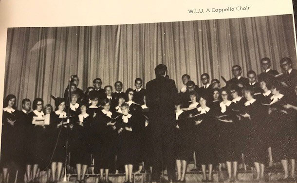 A choir singing in front of a curtain. The conductor has his back to the viewer. This picture is a photograph from a year book; text above the photo says "WLU A Capella Choir"